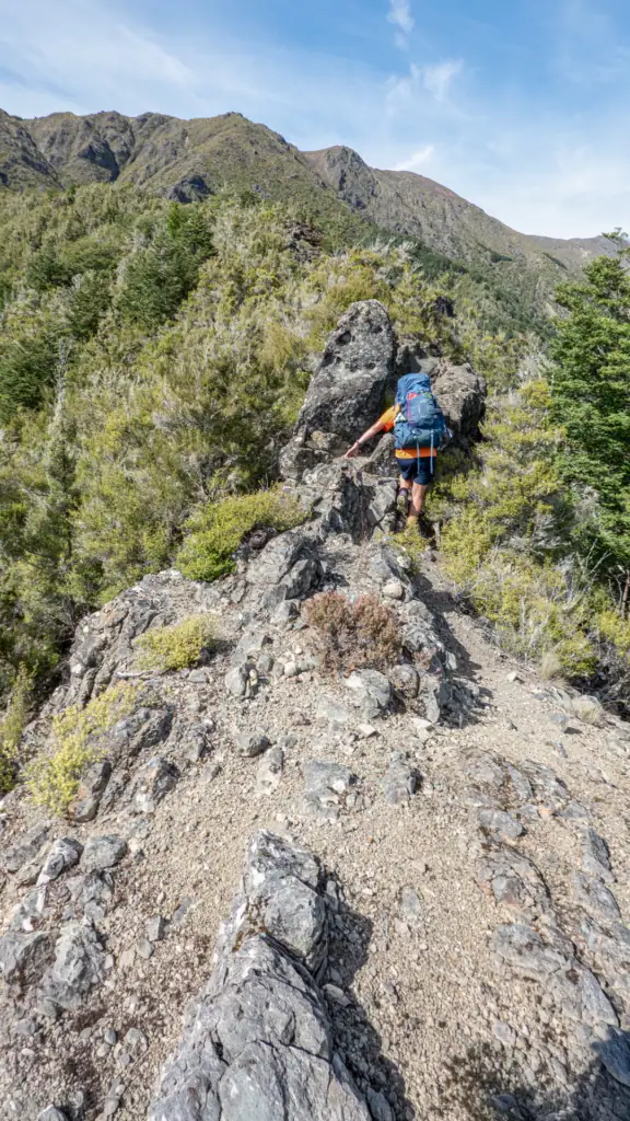 Hiker in orange tshirt with blue pack climbing between rocks up a rugged ridgeline