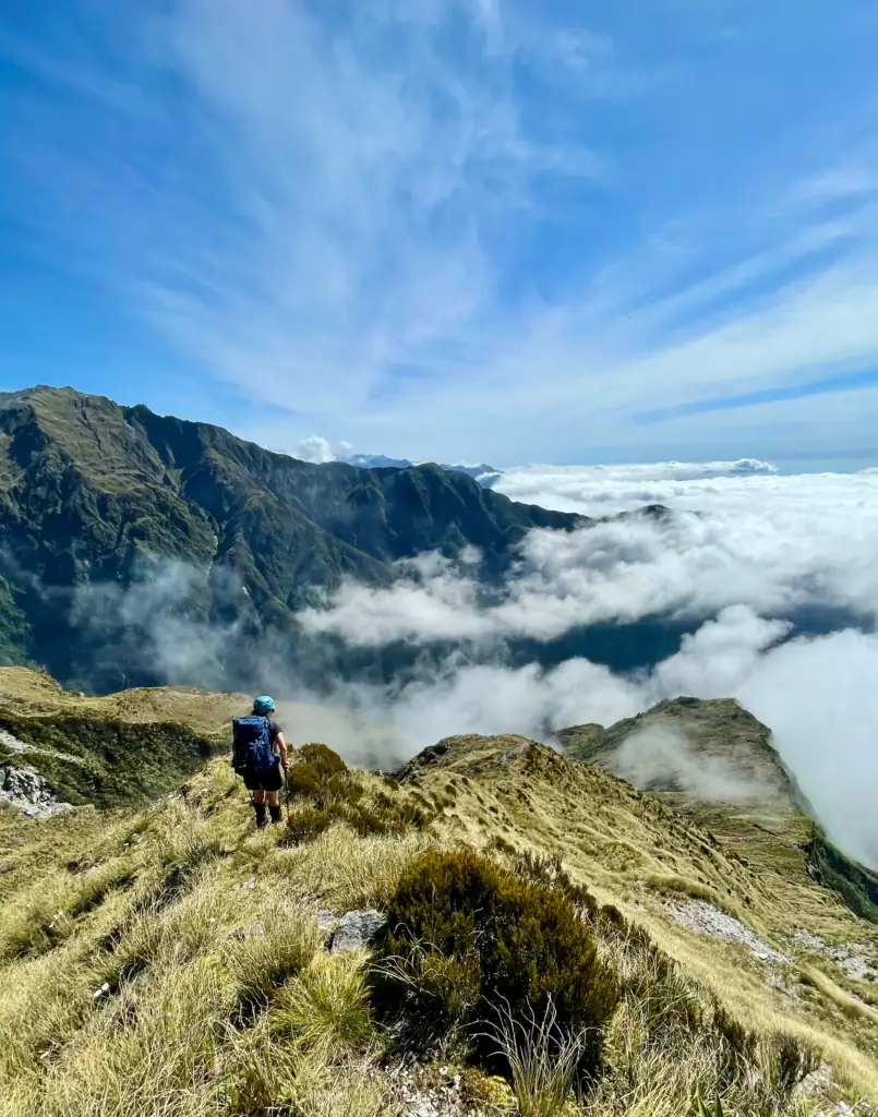 A wide-angle view from behind a hiker descending a grassy, golden-tussock ridge. Wispy clouds cling to the dark green mountain slopes in the distance, and a thick layer of white clouds fills the valley below.
