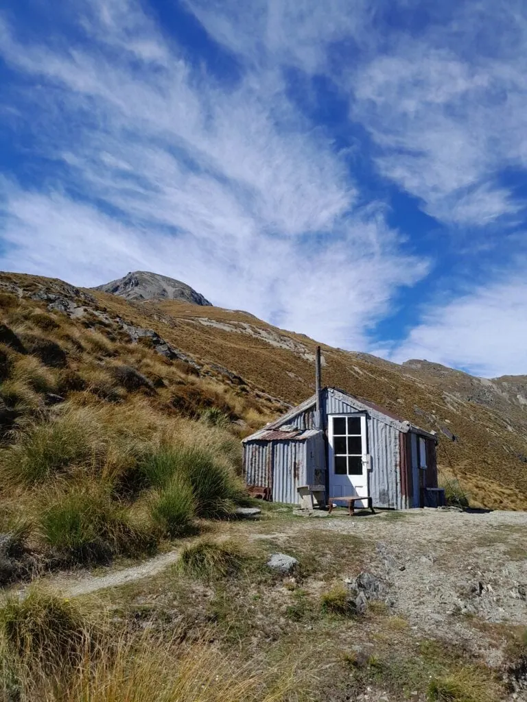 The historic Heather Jock Hut in the Whakaari Conservation Area, with the rocky summit of Mt Alaska (1,965m) rising in the background under a dramatic blue sky