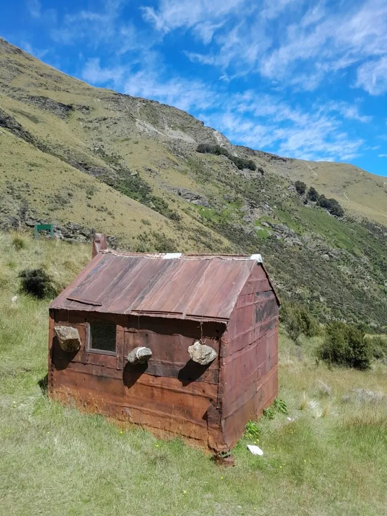 The historic Jean Hut in the Whakaari Conservation Area, showing the unique rusty iron walls and boulders hanging from wires to secure the roof against mountain winds