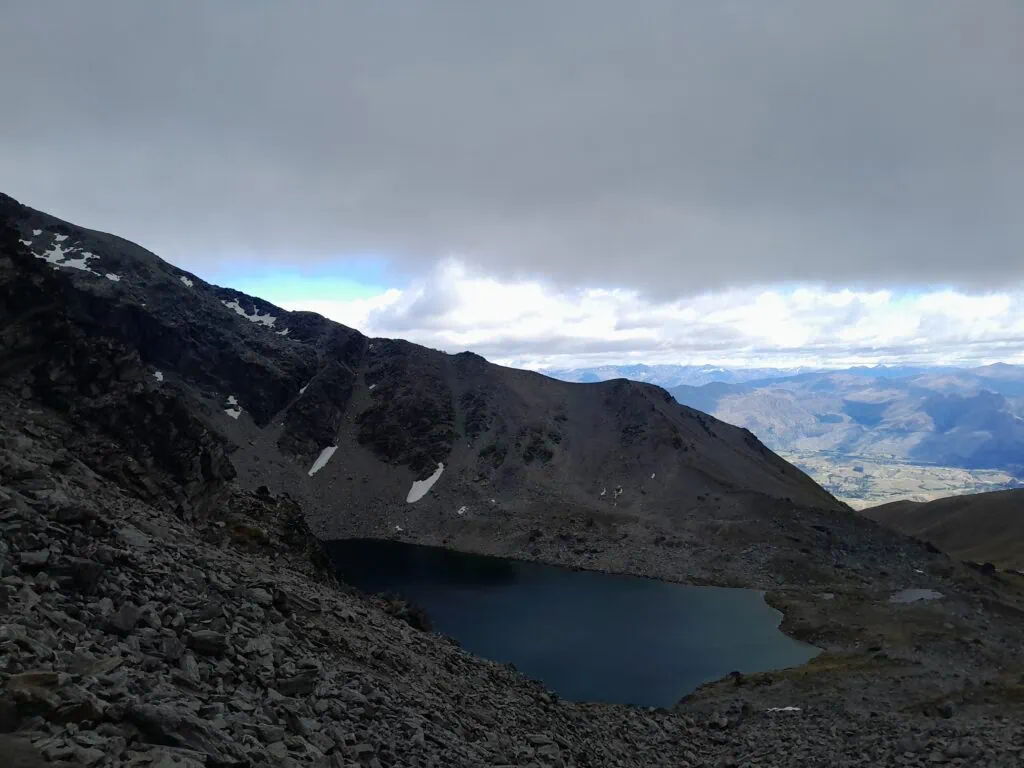 High-angle view of Lake Alta tucked into a rocky glacial cirque in the Remarkables Range, New Zealand, seen from the alpine saddle below Double Cone on the Wye Creek route.