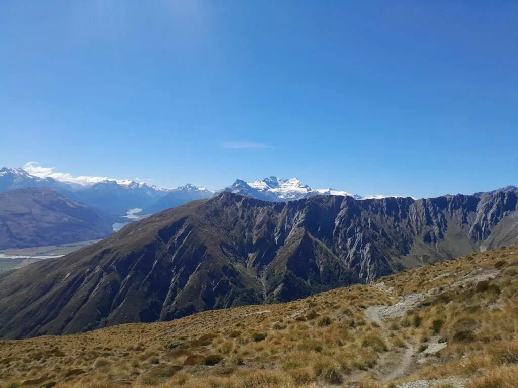 A wide panoramic view of Mount Earnslaw and its glaciers seen from the Long Gully Saddle ridgeline and Southern Alps under a clear blue sky.