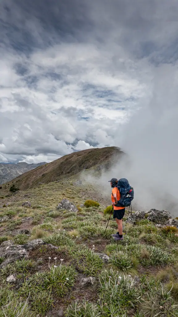 A hiker wearing an orange tshirt with a blue pack stands on a tussocky ridgeline surrounded by cloud