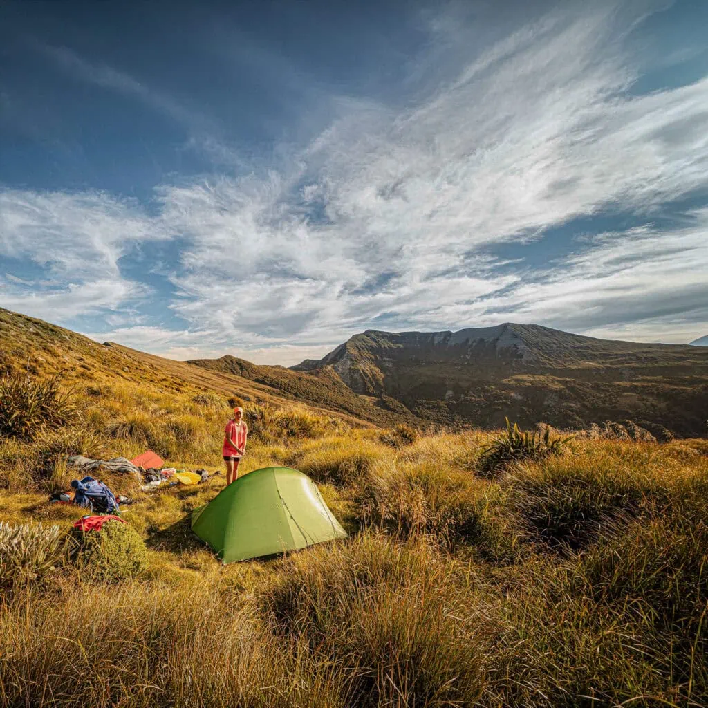 A person camping with a green tent in a lush, mountainous area with dramatic skies. Mudstone formations behind known as Haystack in Matiri Range, Kahurangi National Park