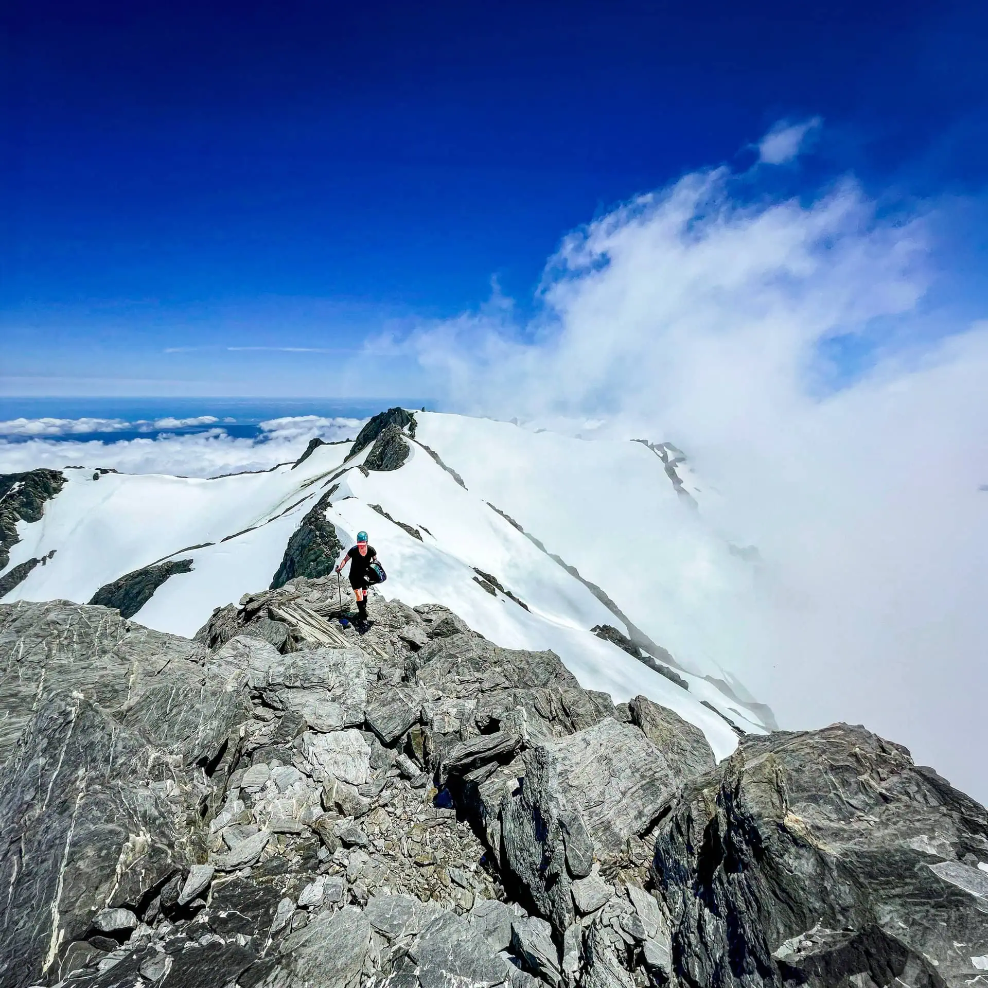 Mountaineer hiking on snow-capped mountain summit with clouds and blue sky.
