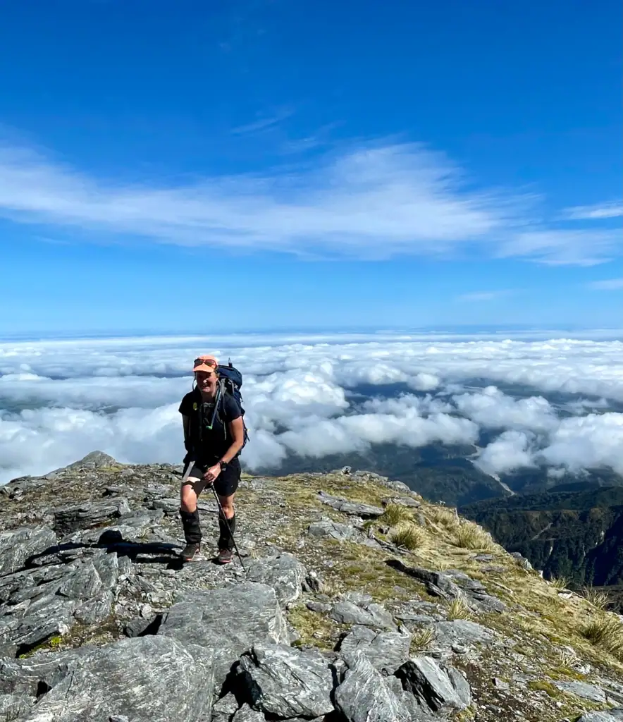 A hiker in an orange cap and backpack stands on a rocky outcrop on the Mt Adams route, smiling. Below them, a vast, flat white cloud inversion stretches to the horizon, making the mountain feel like an island in a sea of clouds.