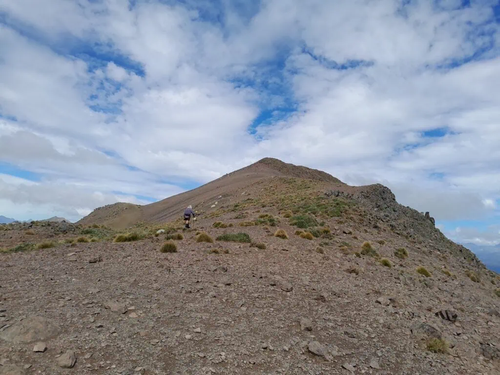 A hiker in a grey top walks along a steep ridgeline covered sporadically in tussock
