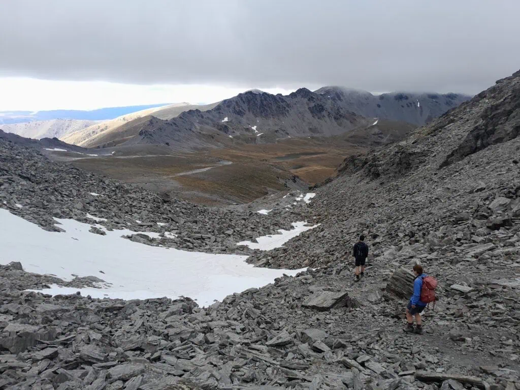 Two hikers the Wye Creek Route, descending a steep scree slope with lingering summer snow patches toward the upper Wye Creek Basin in the Remarkables Range, New Zealand.