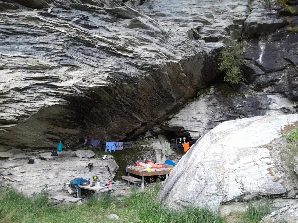 The Earnslaw Burn Rock Biv featuring a wooden sleeping platform, bench, and clothesline with tramping gear under a large overhanging rock in the Southern Alps.