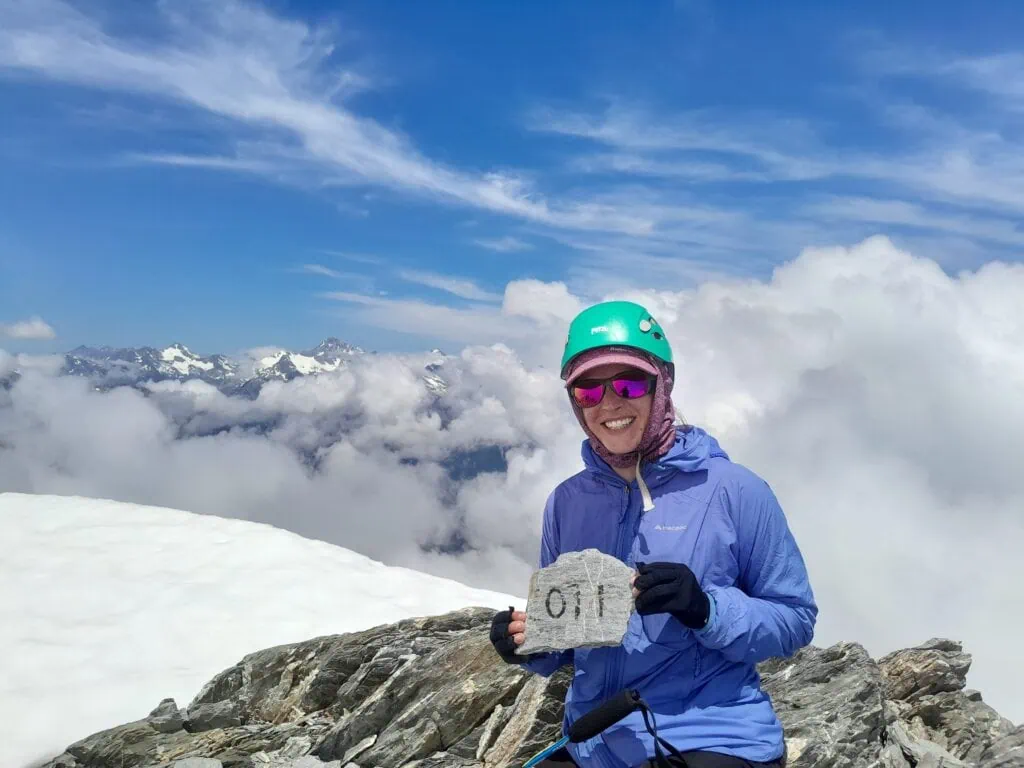 A hiker in a blue helmet and blue top stands smiling on the summit of Mt Adams holding an engraved rock with the southern alps in view behind her