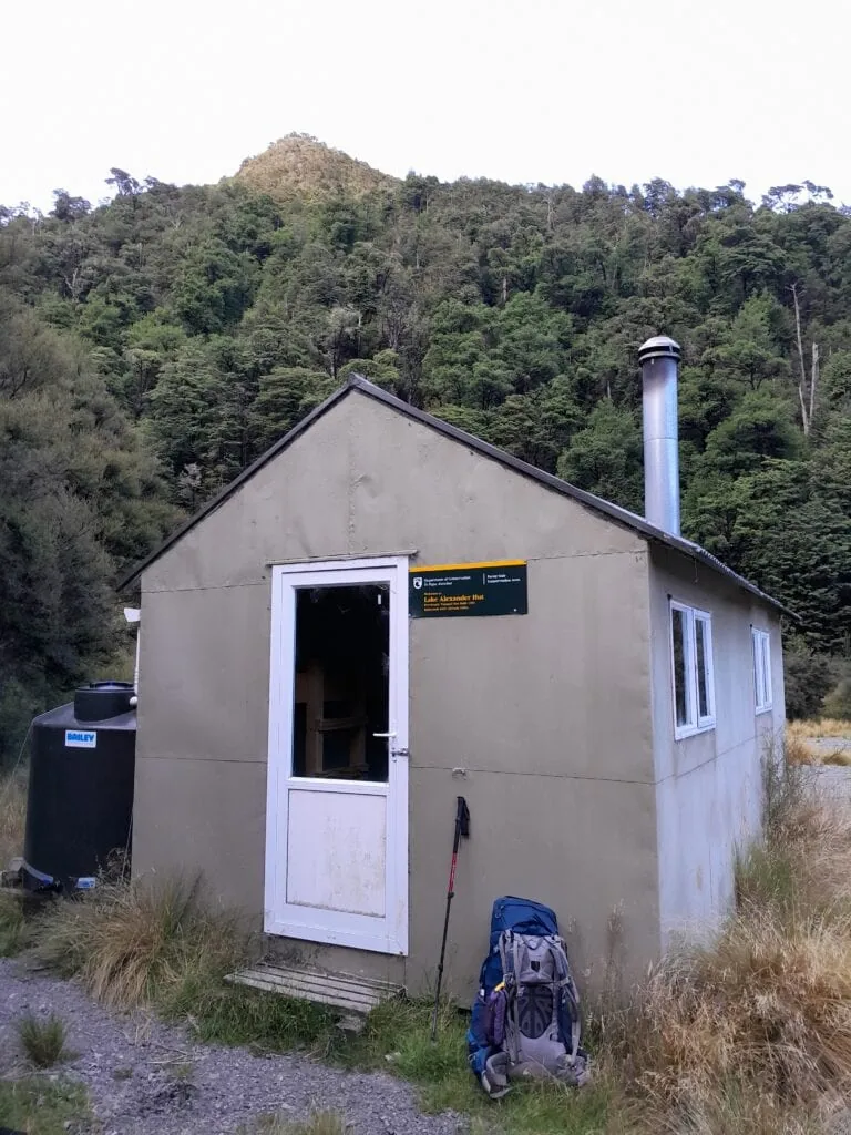 A department of conservation hut called Lake Alexander hut is surrounded by trees and tussock with a hiker's pack in front of it