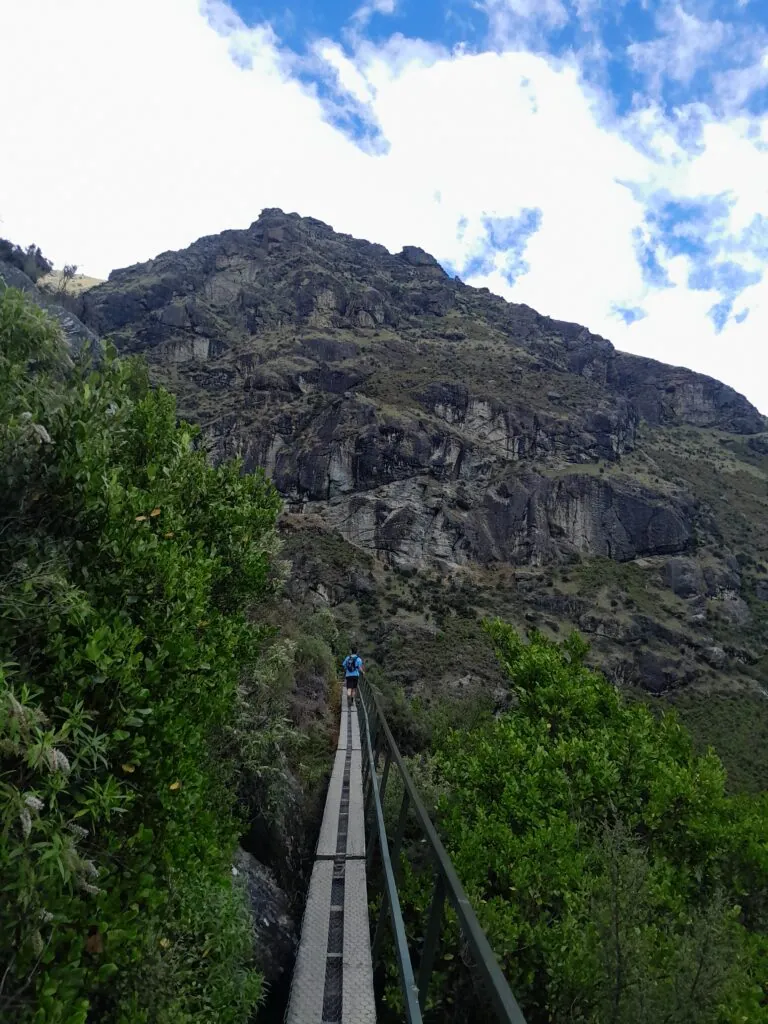 A hiker walking along the narrow platform of the Wye Creek hydro dam walkway, passing beneath towering schist rock bluffs in the Remarkables Range on the Lower Wye Creek Route.