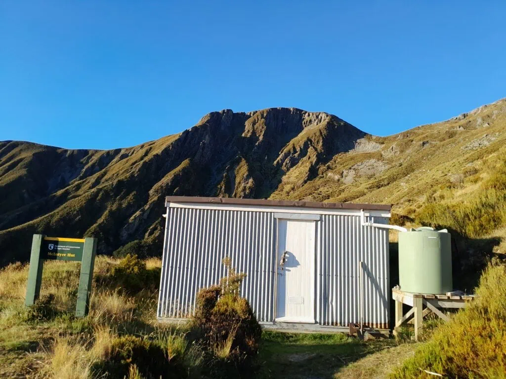 Exterior of the historic McIntyre Hut in the Whakaari Conservation Area, featuring its corrugated iron walls, green water tank, and a backdrop of rugged alpine ridges near Glenorchy