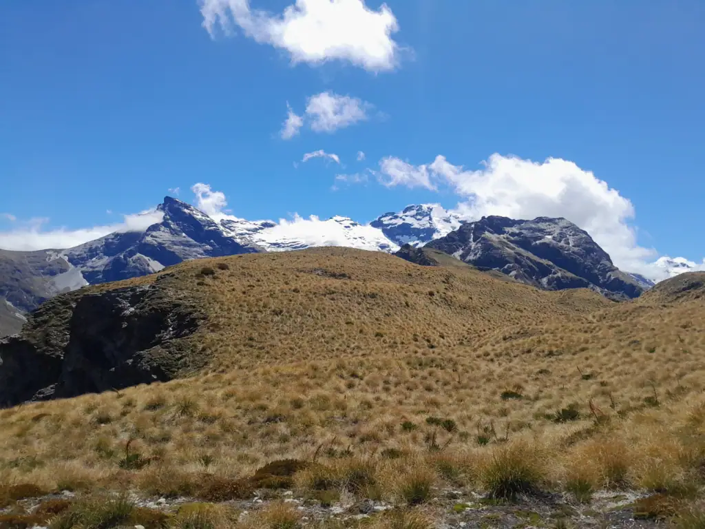 A panoramic view of Mount Earnslaw (Pikirakatahi) and its glaciers from a high alpine ridgeline in the Southern Alps, New Zealand, featuring golden tussock and clear blue skies.