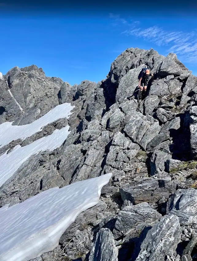 A climber wearing a backpack and orange hat scrambles down a steep, craggy rock face on the Mt Adams route. Patches of bright white snow are nestled in the hollows of the grey, fractured rock under a clear blue sky.