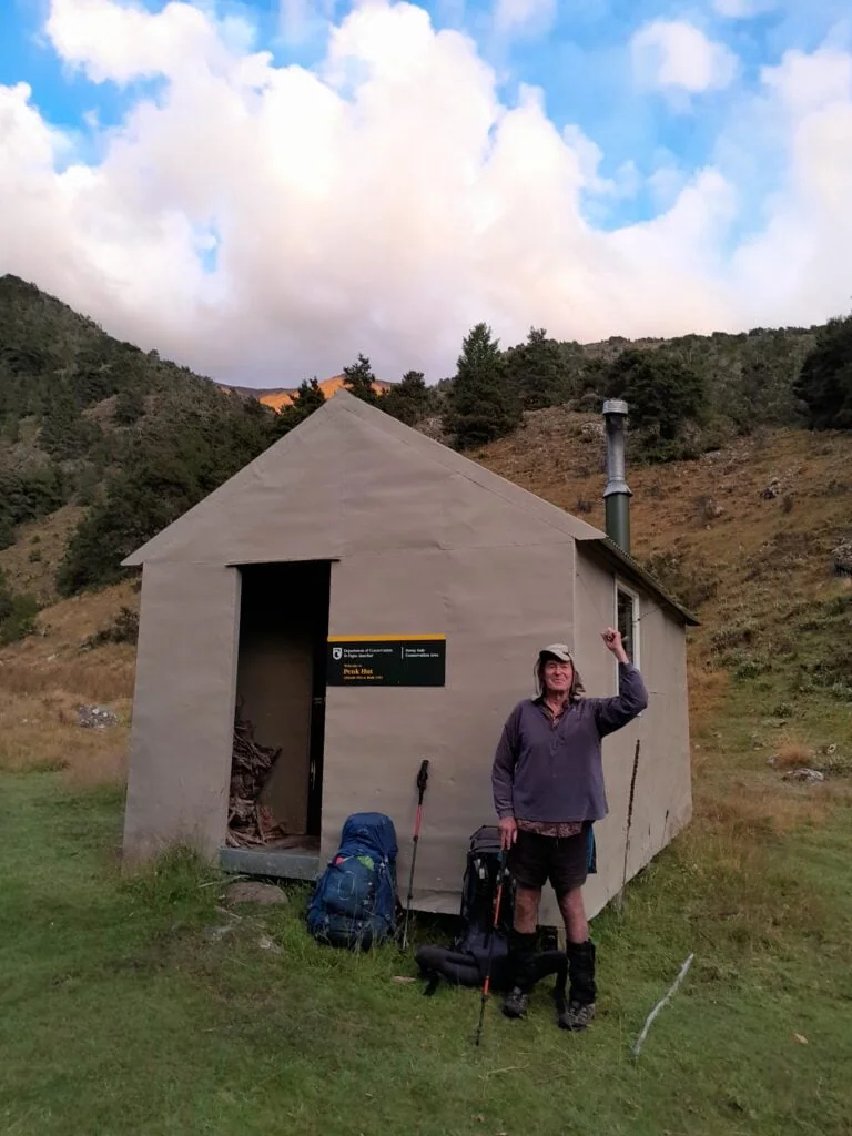 A happy hiker wearing a grey top at a hut called penk hut with a blue hiking pack next to him.