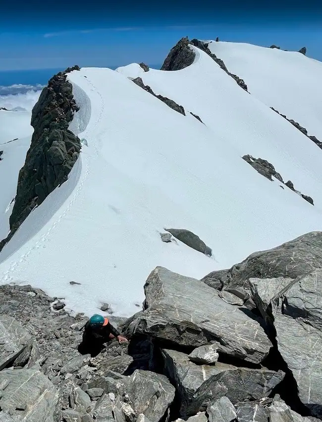 A high-angle shot looking down at a climber navigating a steep field of grey boulders. In the background, a pristine, undulating snow ridge leads upward against a deep blue sky.