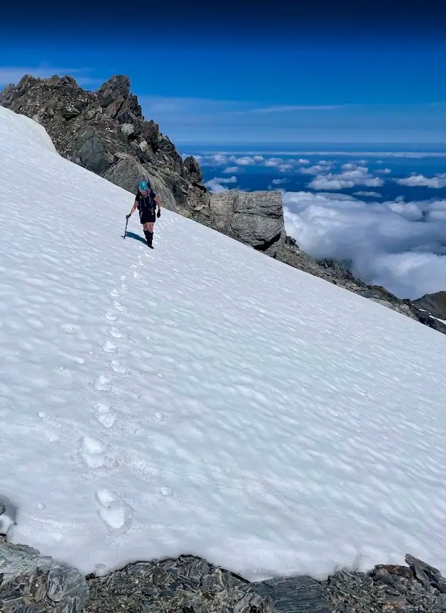 A climber uses an ice axe to traverse a steep, snowy slope on Mt. Adams. A trail of footprints marks their path across the white expanse, with a dramatic drop-off into a cloud-filled valley on the right.