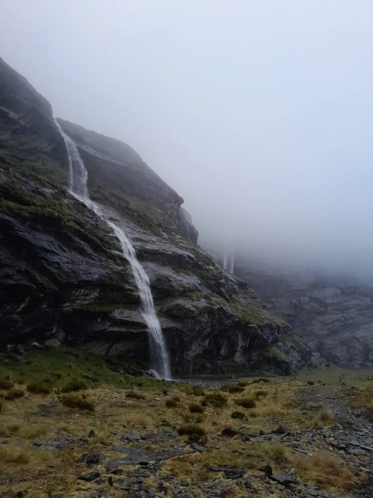 A tall alpine waterfall cascading down a dark rock face into the head of the Earnslaw Burn valley, partially obscured by thick mountain mist and clag