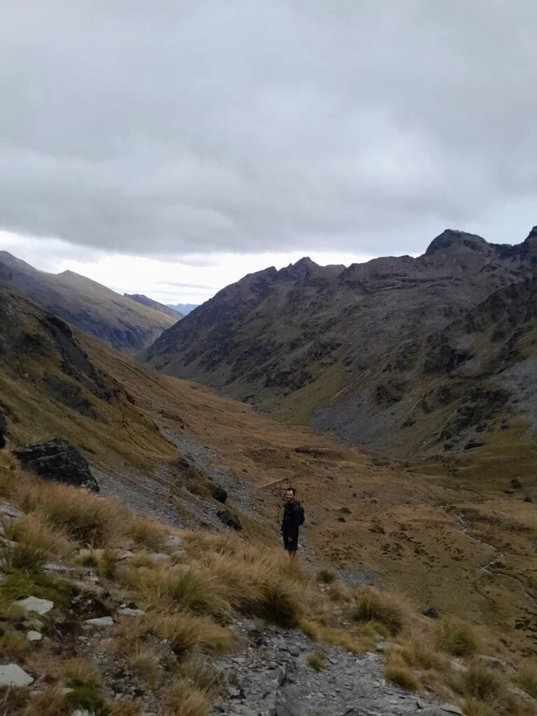 A hiker standing on a rocky trail overlooking the vast, golden Wye Creek Basin surrounded by the steep, dark ridges of the Remarkables Range in New Zealand.