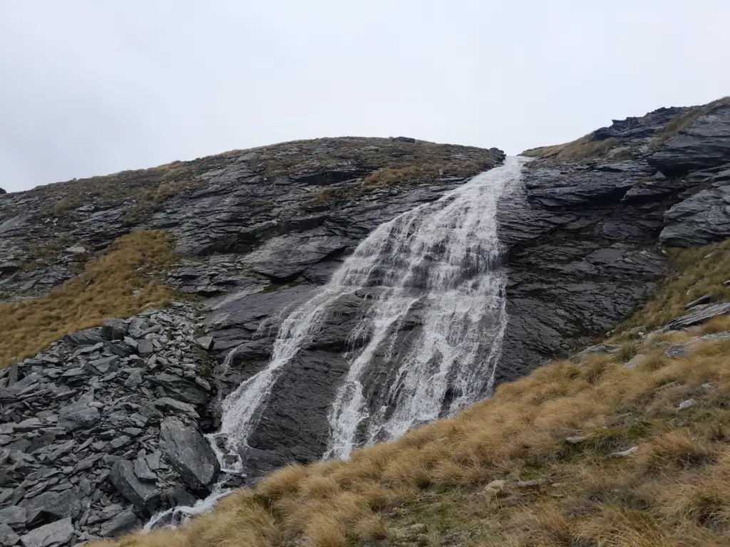 A dramatic alpine waterfall cascading down steep dark rock faces into the Wye Creek Basin, located in the Remarkables Range on the Wye Creek Route in New Zealand.