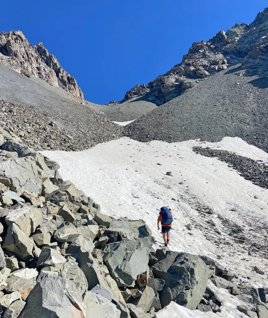 A tramper with a large backpack ascending a permanent snow patch below Alma Col (2,139m) in the Two Thumb Range. The technical off-track route involves navigating steep loose rock and alpine snow in Canterbury, New Zealand.