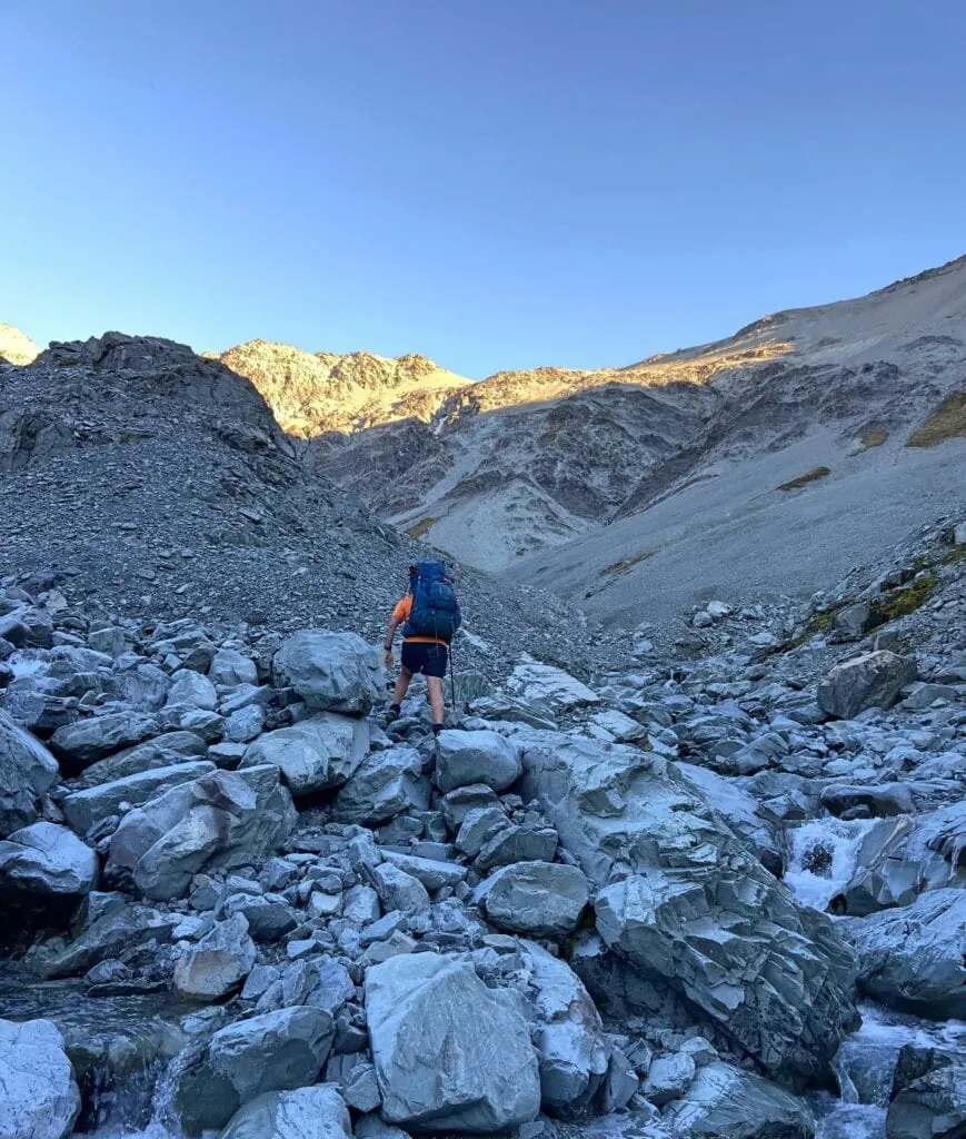 A tramper with a large backpack navigating a field of massive boulders while en route to Balaclava Saddle. The image showcases the challenging off-track alpine terrain of the Two Thumb Range in Canterbury, New Zealand.