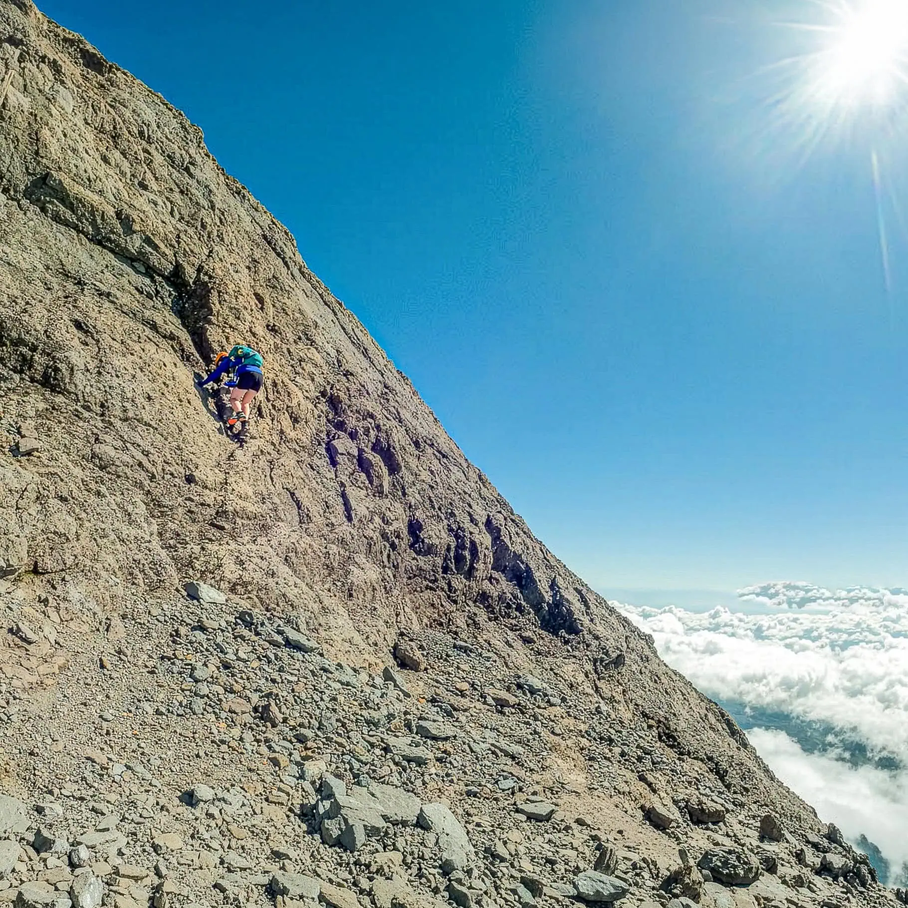 A climber scrambles up a step on the East Ridge above the clouds and under a clear blue sky