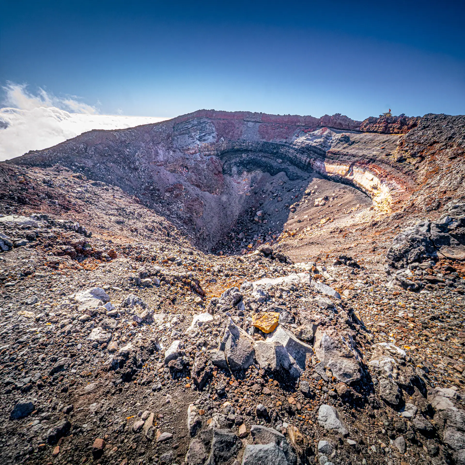 A wide-angle, high-angle shot looking into the deep, rocky crater of Mt. Ngauruhoe under a clear blue sky. The crater walls show layers of red and grey volcanic rock, with a field of jagged boulders and loose scree in the foreground.