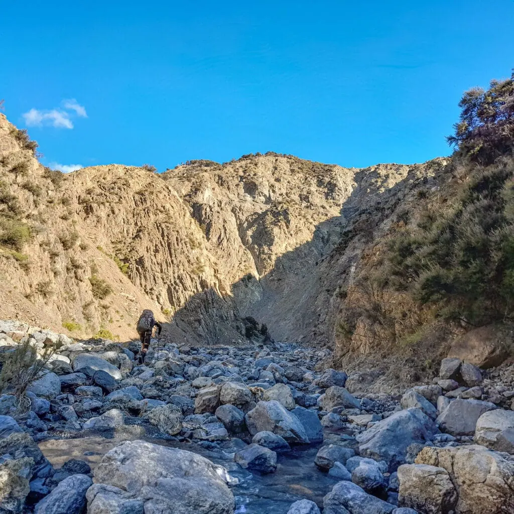 A hiker with a black pack walks up a boulder covered Doddemeade Stream with tall and eroded gorge walls in Seaward Kaikōura Range, Ka Whata Tu o Rakihouia Clarence