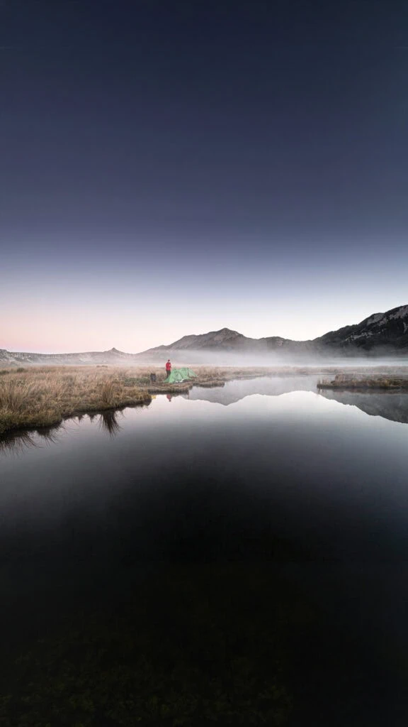 A hiker in a red jacket stands next to a green tent and a tarn with their reflection in it surrounded by a think layer of early morning mist with the mudstone peaks Haystack and Needle in the background in Kahurangi National Park