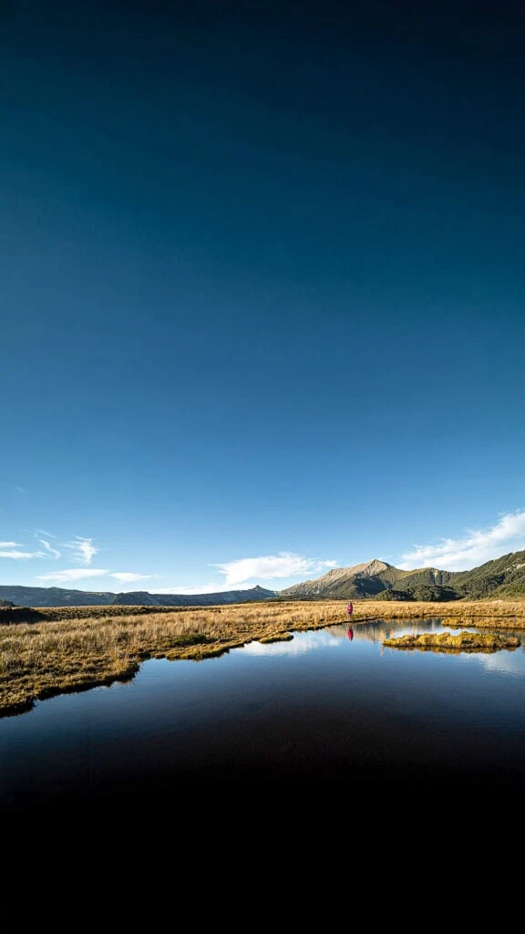 A hiker stands to a tarn with their reflection in it under a bright blue sky with two mudstone peak formations in the background, the Needle and the Haystack, in the Matriri Range