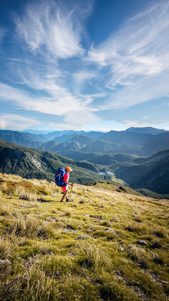 Hiker in pinnk tshirt with orange cap and large blue pack descending over tussock and grass covered slopes to Hurricane Hut with a lake called Lake Jeanette nestled amongst the bush in the background