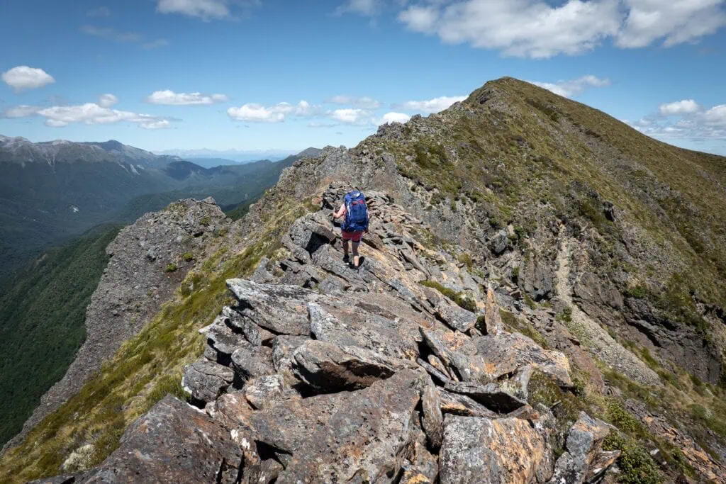 Hiker with a large blue pack navigating across large rugged boulders on a narrow ridgeline on the Matiri Range in Kahurangi National Park
