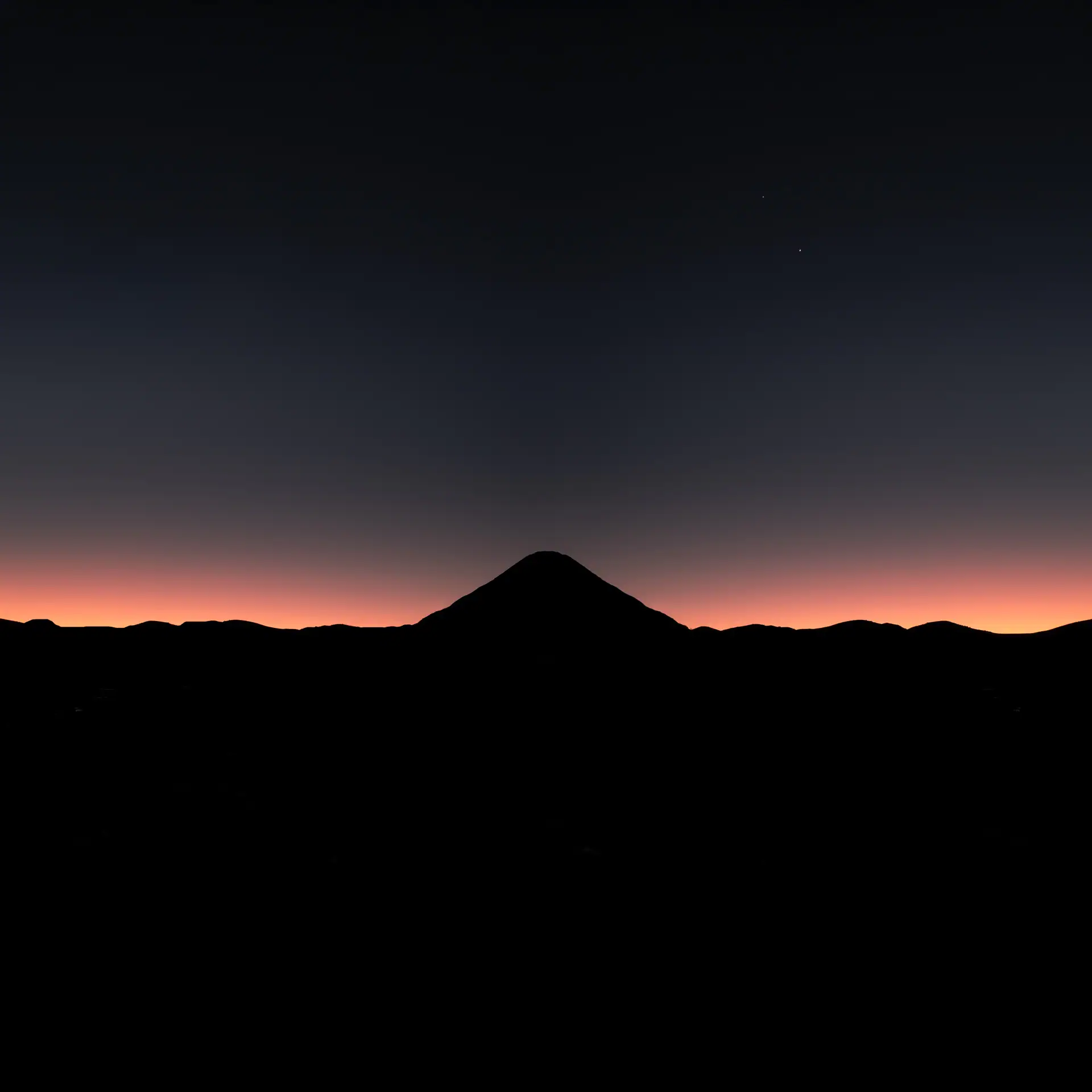 A dark silhouette of the conical Mt Ngauruhoe centered against a vibrant orange and deep blue dawn horizon, viewed from a distance across a black foreground.