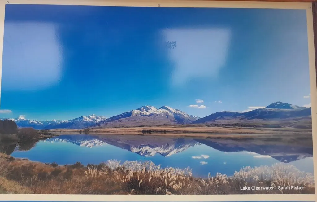 A scenic photo of Lake Clearwater with a snowy Mt Poots, Cloudy Peak Range and Two Thumb Range in Hakatere Conservation Area featured on a local information sign in Mount Somers Village