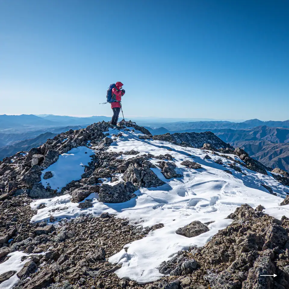 Hiker on the summit of a rocky peak in the Raglan Range NZ with snow on the ground and a clear blue sky