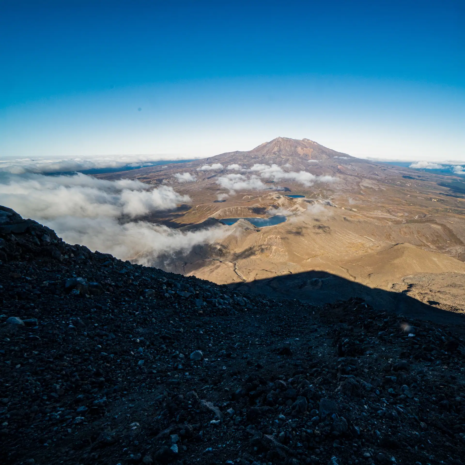 An expansive view from the rocky volcanic slope of Mt Ngauruhoe looking across the blue Tama Lakes toward the massive, peak of Mt Ruapehu under a clear sky.