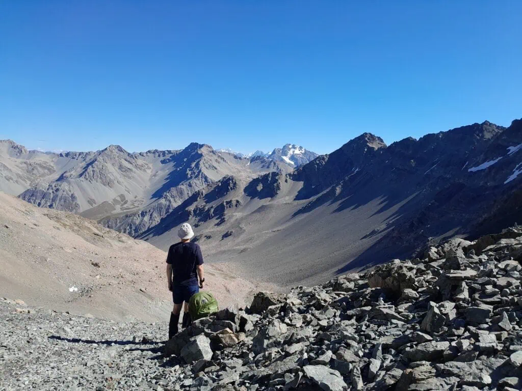 A tramper standing at Alma Col (2,139m) overlooking the glaciated peaks of the Southern Alps including Aoraki/Mt Cook, Mt Sibbald and Exeter Peak. The rugged alpine scenery of the Two Thumb Range, Canterbury, is visible under a clear blue sky.