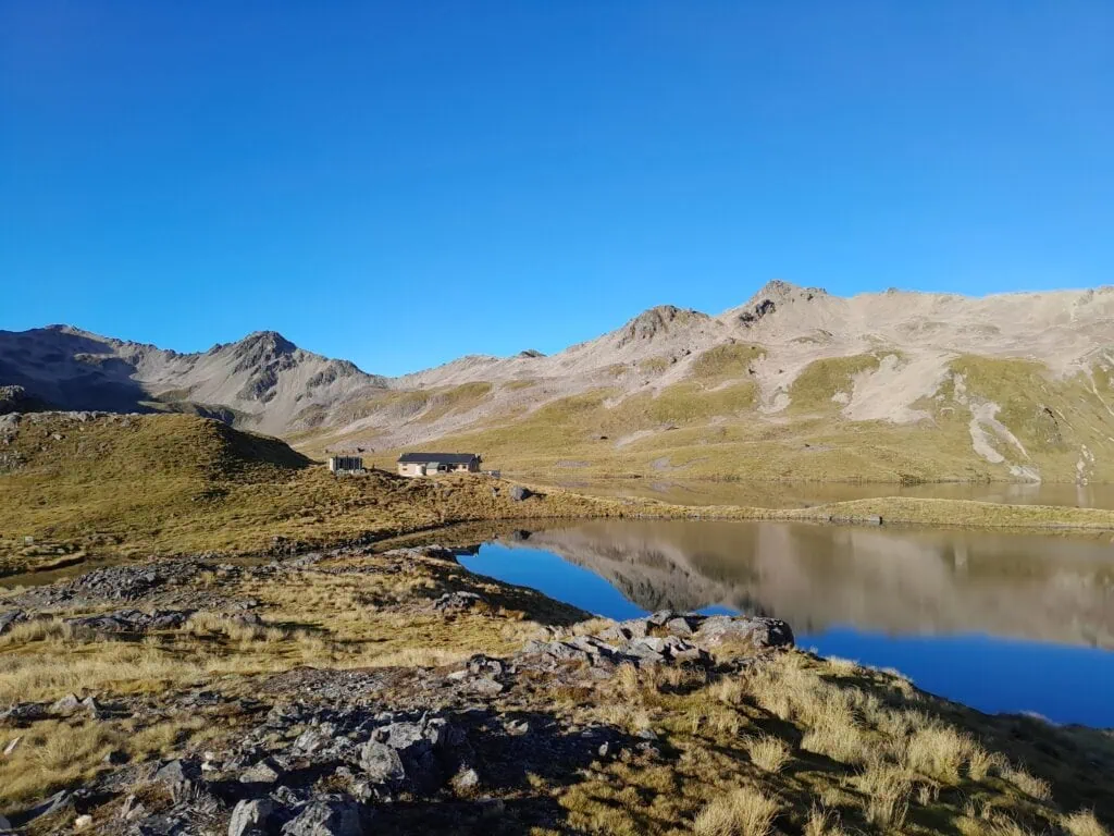 A blue bird day with a large tarn with reflections in it and Angelus Hut