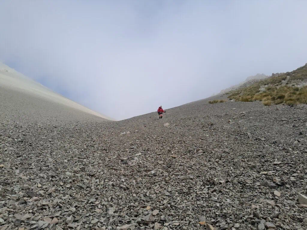 A tramper in a red jacket descending a vast grey scree slope from Brabazon Saddle in the Two Thumb Range, Canterbury. The high-altitude landscape is partially obscured by mountain mist and low cloud.