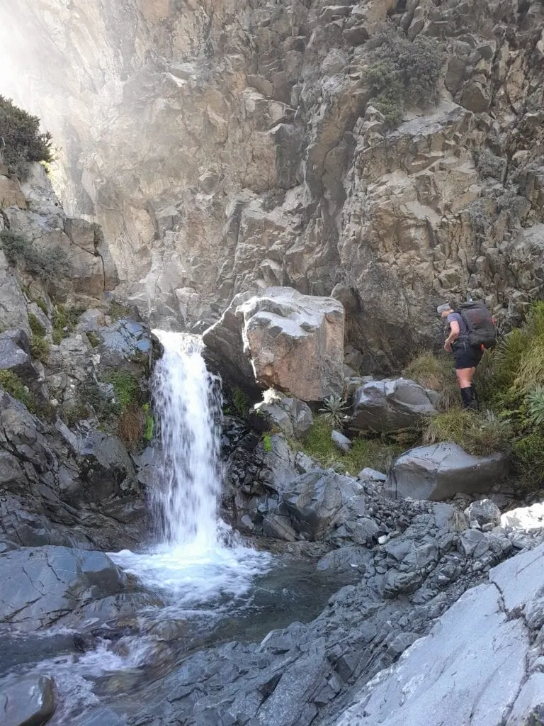 A tramper standing next to a mountain waterfall in a narrow rock gorge at Black Birch Creek, Two Thumb Range, during the climb to Inkerman Saddle. The rugged alpine terrain features steep grey bluffs and technical off-track navigation in the Canterbury high country.
