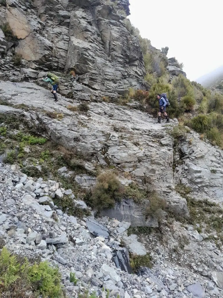 Trampers navigating rocky bluffs and steep terrain above Black Birch Creek in the Two Thumb Range, Canterbury. Two hikers with packs are shown sidling across a grey rock face in a rugged alpine environment.