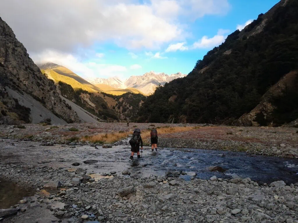 Three trampers crossing the shallow, rocky Bush Stream and heading up the valley on the Te Araroa Trail section of the Two Thumb Range, Canterbury high country. Crooked spur and Rugged mountains and scree slopes are illuminated by early morning sun in the distance