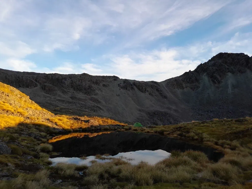 Early morning sun slight on a basin next to a tarn with a green camping tent under Robert Ridge