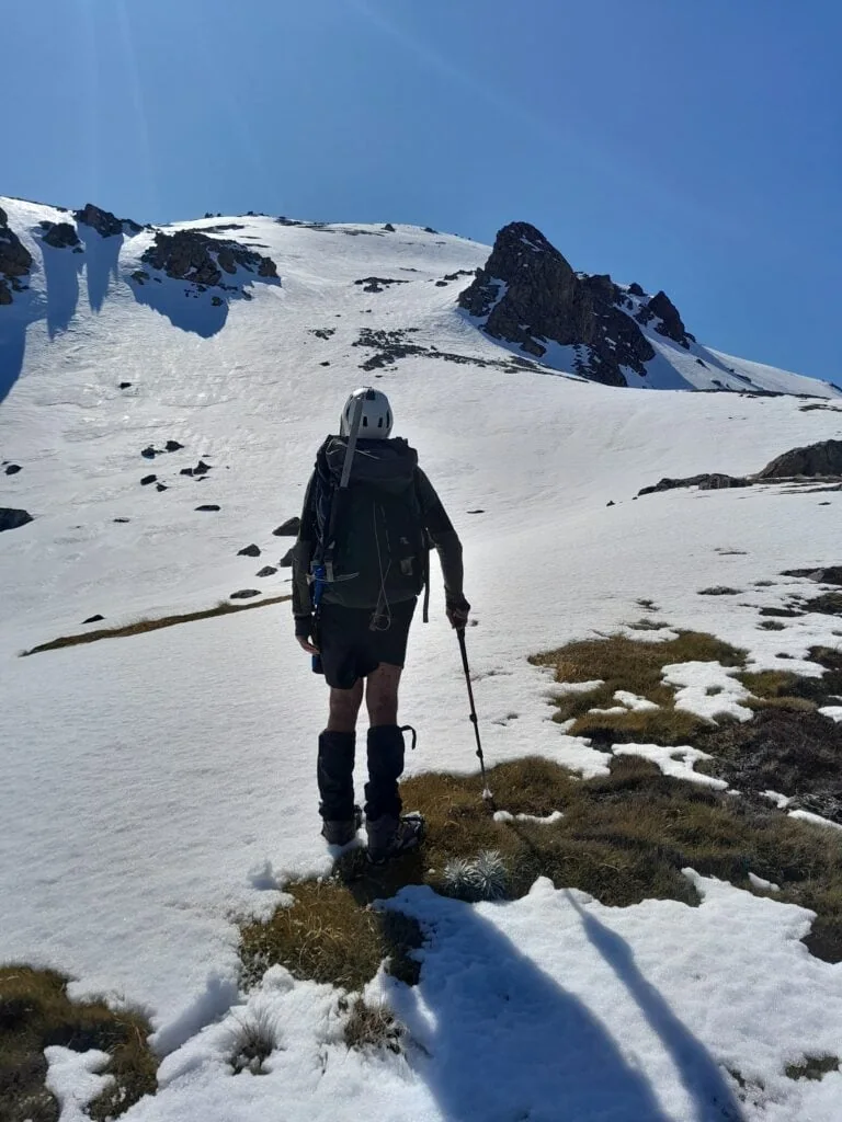 A hiker with alpine gear stands at the snowline looking up a steep slope on the way to Princess Bath in St James Conservation Area