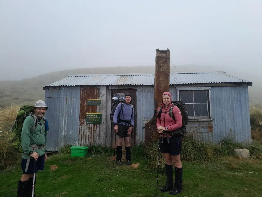 Three trampers stand outside an old musterers' hut known as Crooked Spur Hut on the Two Thumb Range section of the Te Araroa Trail. They are surrounded by mist.