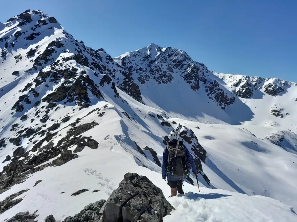 A hiker in alpine gear stands in deep soft snow on the ridge to Mt Princess with a snow-covered frozen Princess Bath tarn on his right, St James Conservation Area