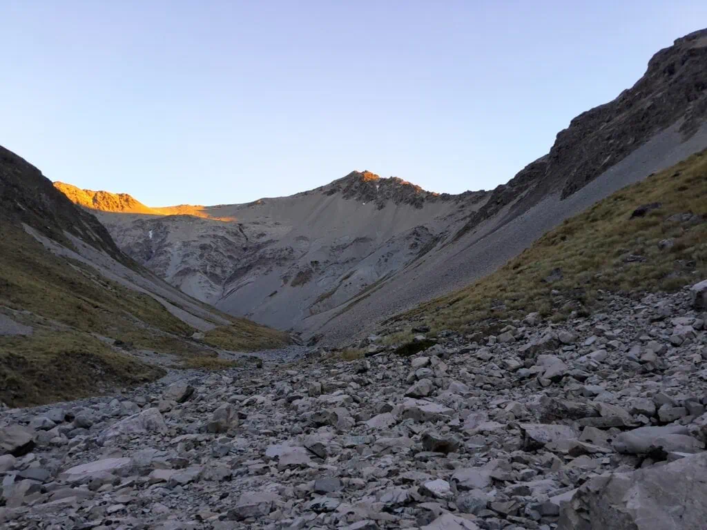 Early morning view of the rocky upper catchment of North East Gorge Stream, looking toward Balaclava Saddle in the Two Thumb Range. The rugged alpine terrain features a boulder-filled riverbed with sunrise light hitting the high peaks including Graf Spee
