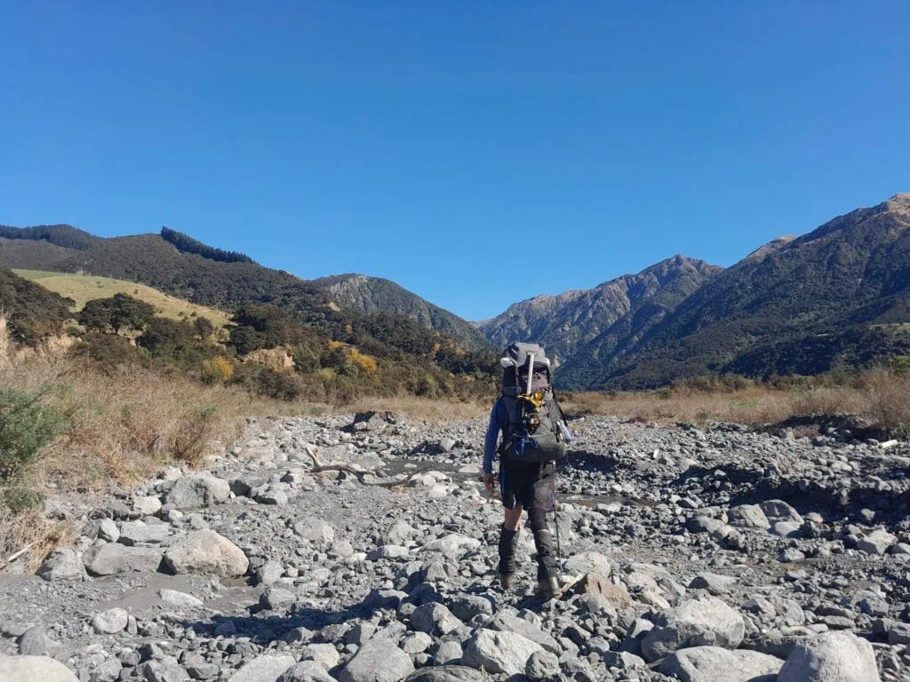 A hiker with alpine gear on a large pack walks through boulders in George Stream on the way to Mt Tarahaka in Seaward Kaikōura Range in Ka Whata Tu o Rakihouia Conservation Park